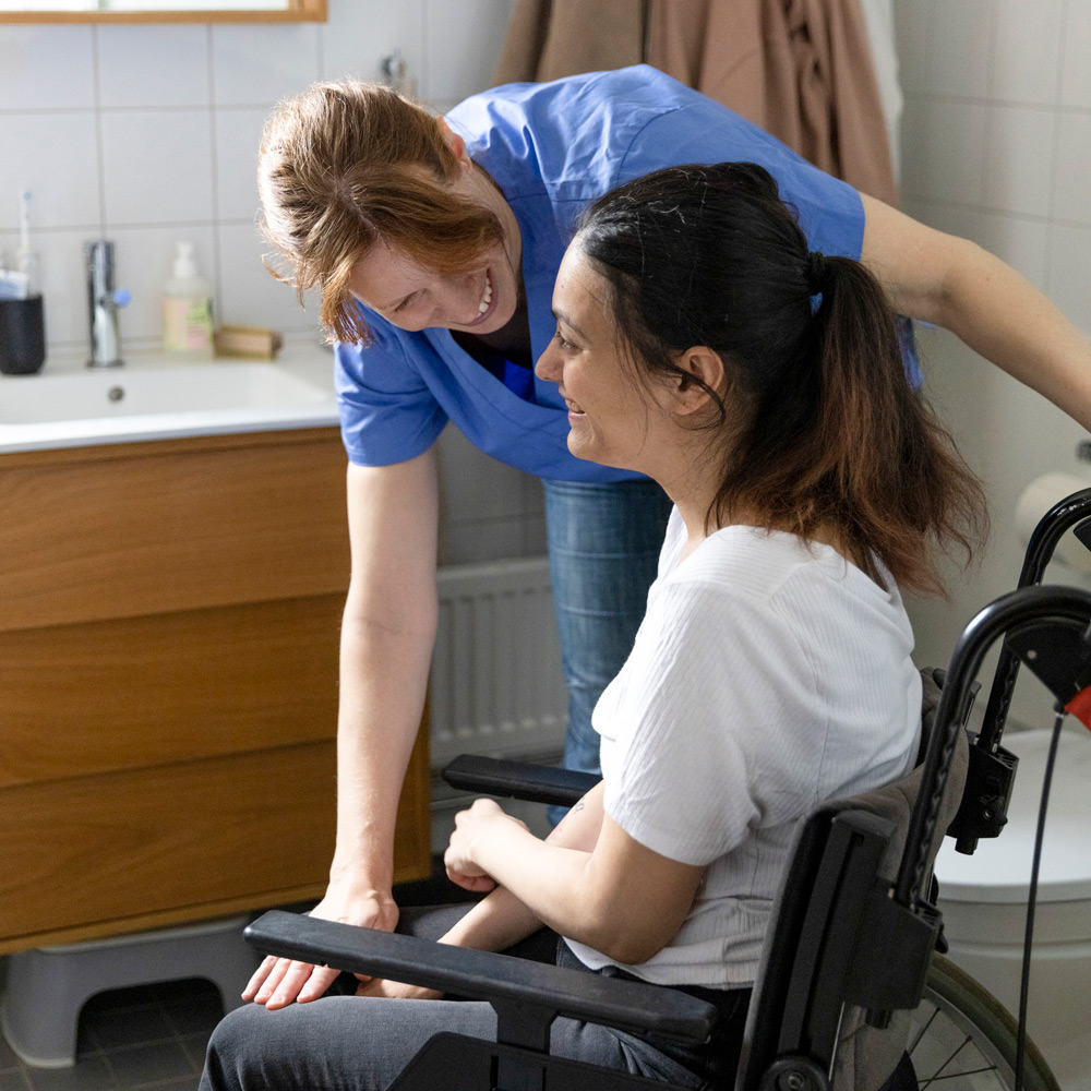 En glad kvinnlig vårdgivare i blå sjuksköterskeuniform böjer sig ned och pratar engagerat med en ung kvinna med paraplegi som sitter i en rullstol i ett badrum hemma.